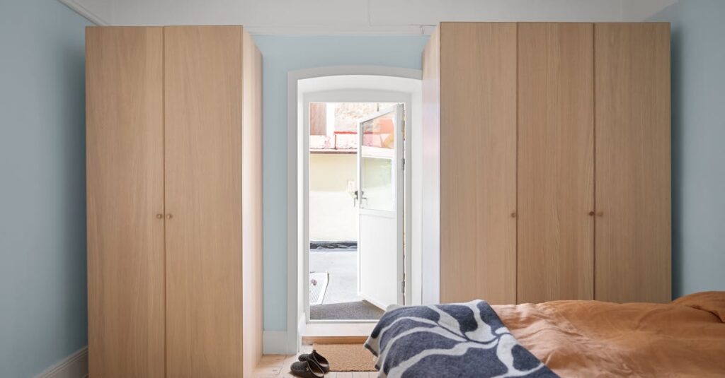 A minimalist bedroom with sunlight streaming through a door, featuring wooden wardrobes and a cozy bed with colorful blankets.